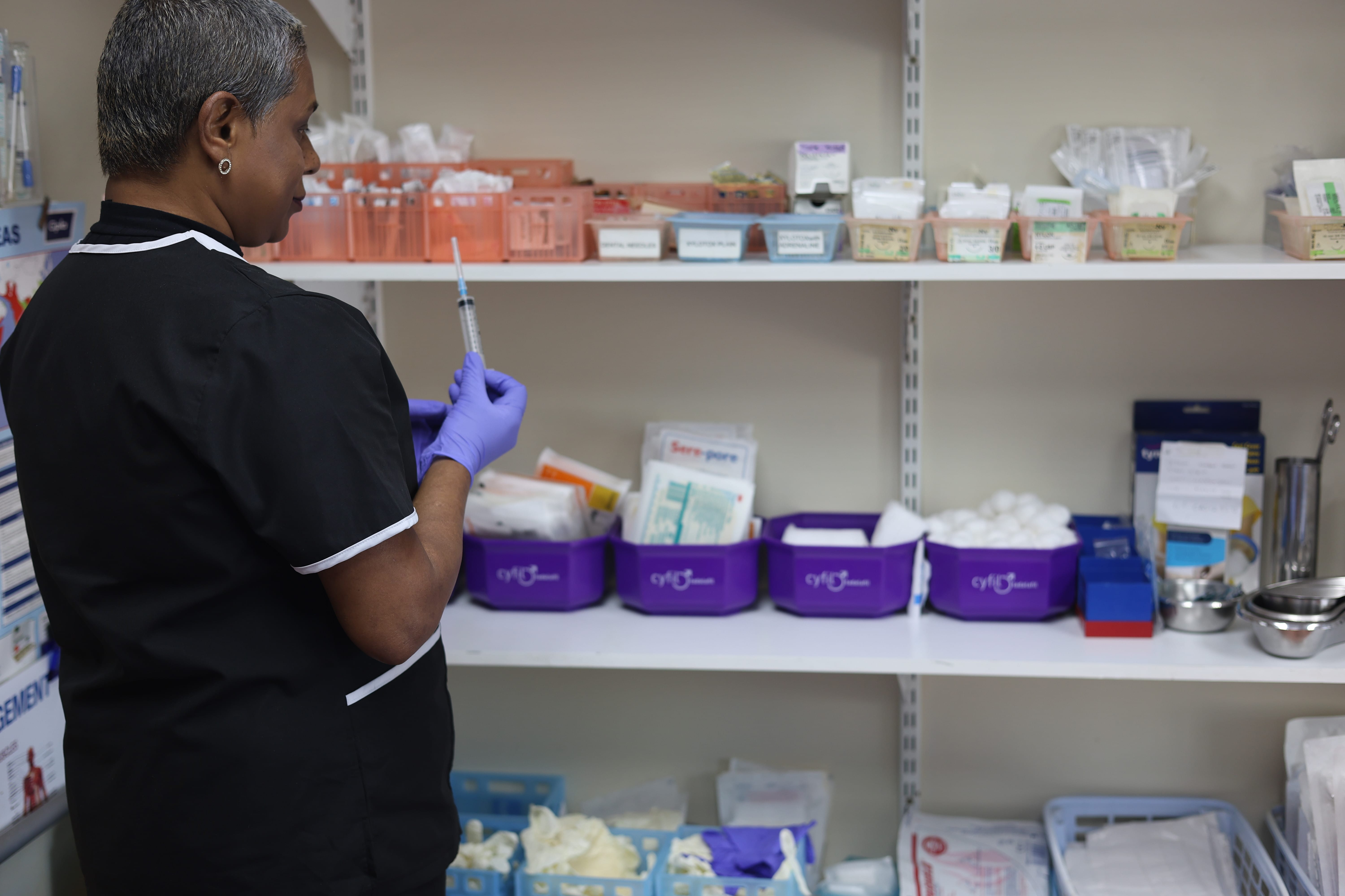 A clinician preparing a syringe beside shelves of supplies at Sunningdale Medical Centre